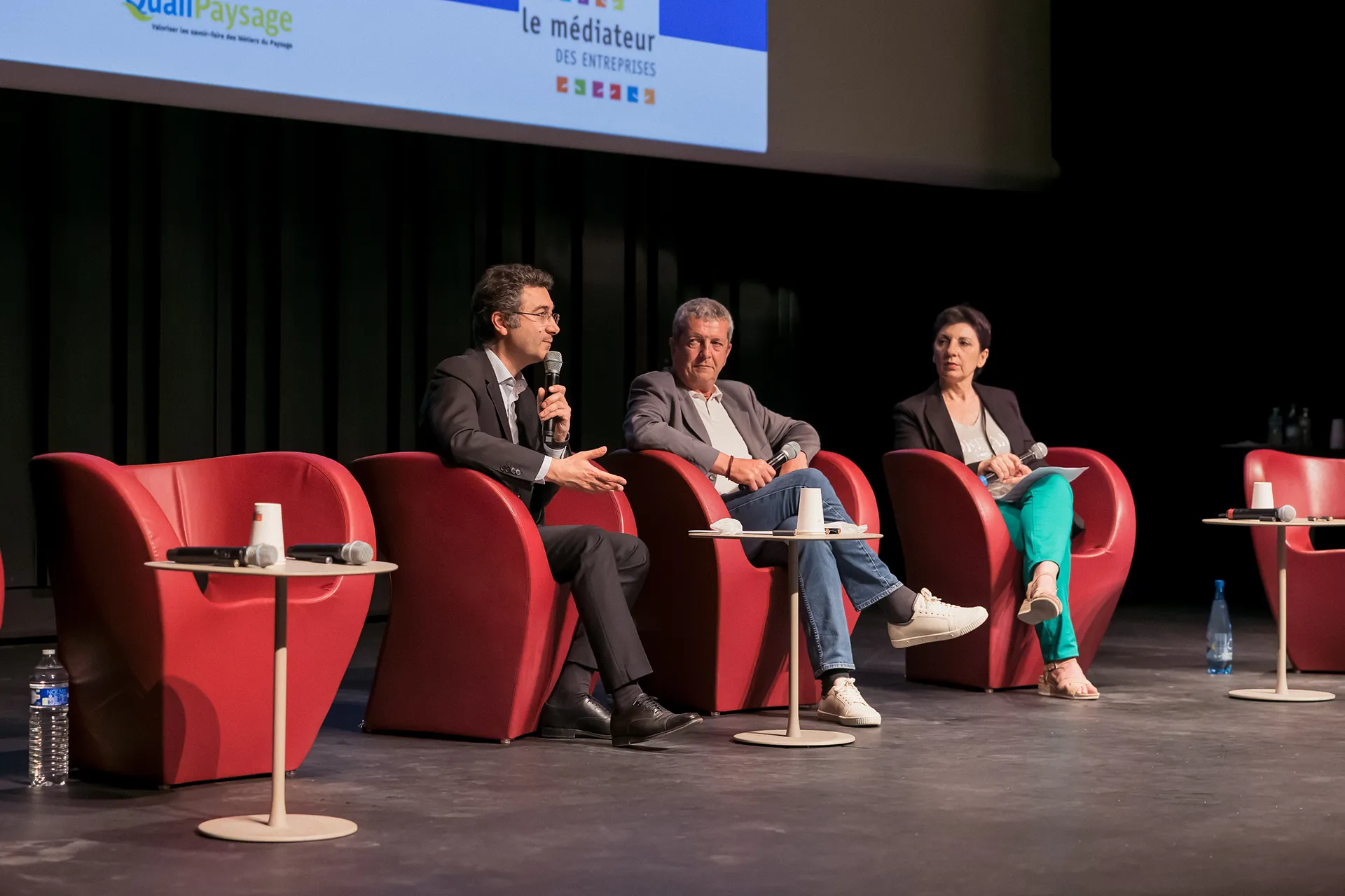 Trois intervenants assis dans des fauteuils rouges sur une scène de conférence. L’écran au-dessus d’eux porte les logos QualiPaysage et Médiateur des entreprises.